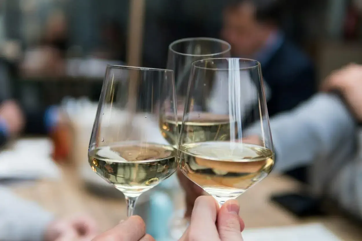 Drei Gläser mit hellem Wein, ein Sauvignon Blanc trocken, werden zum Anstoßen gehalten. Im Hintergrund verschwimmen die Silhouetten von Menschen.