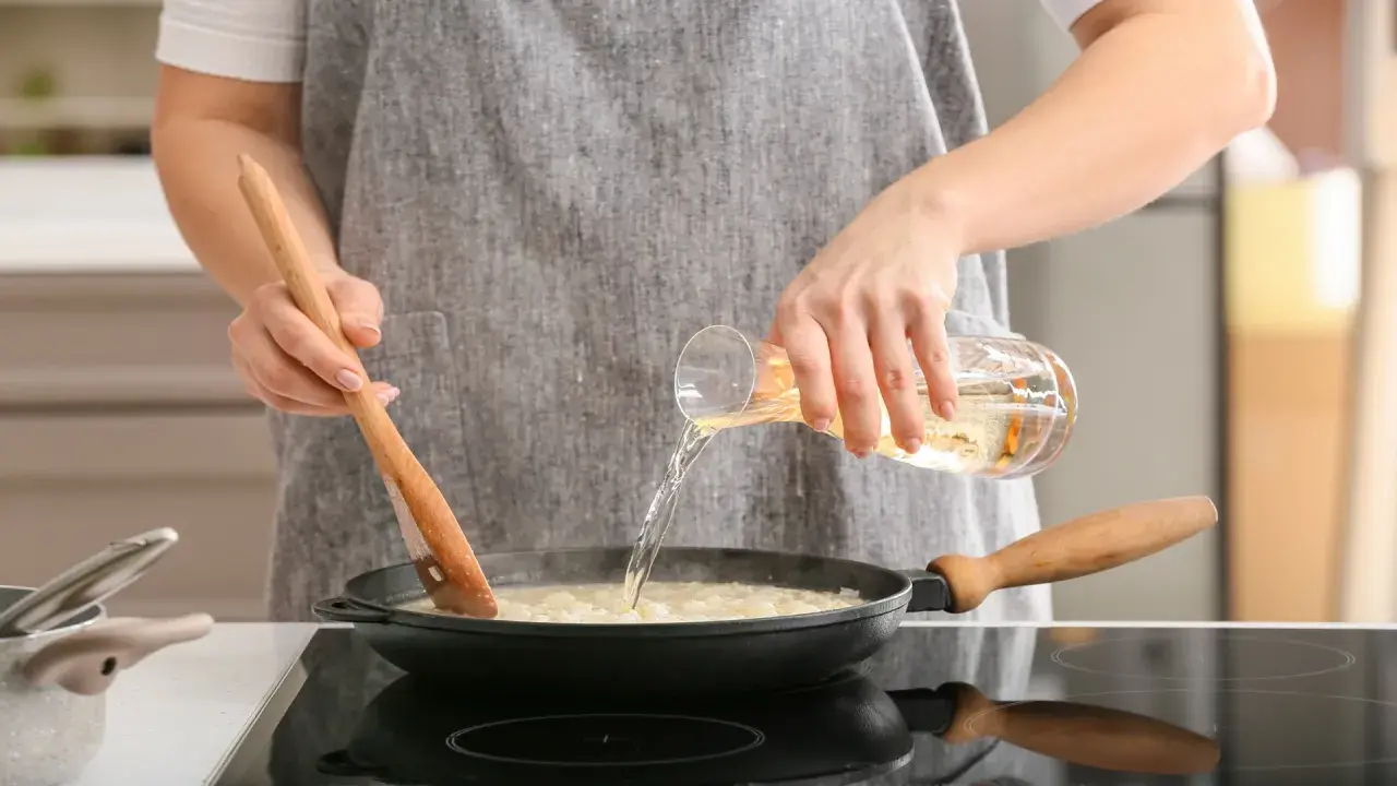 Eine Person verkocht Alkohol beim Kochen. Sie gießt eine Flüssigkeit aus einem Glas in eine Pfanne, während sie mit einem Holzlöffel umrührt.