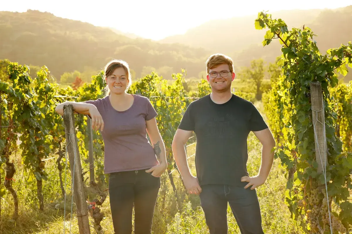 Zwei junge Winzer aus der Pfalz lächeln in der goldenen Abendsonne inmitten ihrer Weinreben.