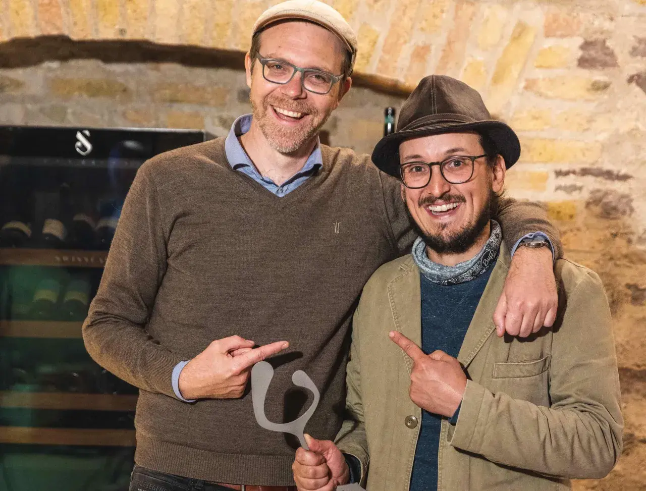Zwei Männer mit Hüten und Brillen halten eine graue Skulptur. Sie zeigen darauf und lächeln. Ein Zeichen für "Wein gegen Rassismus".