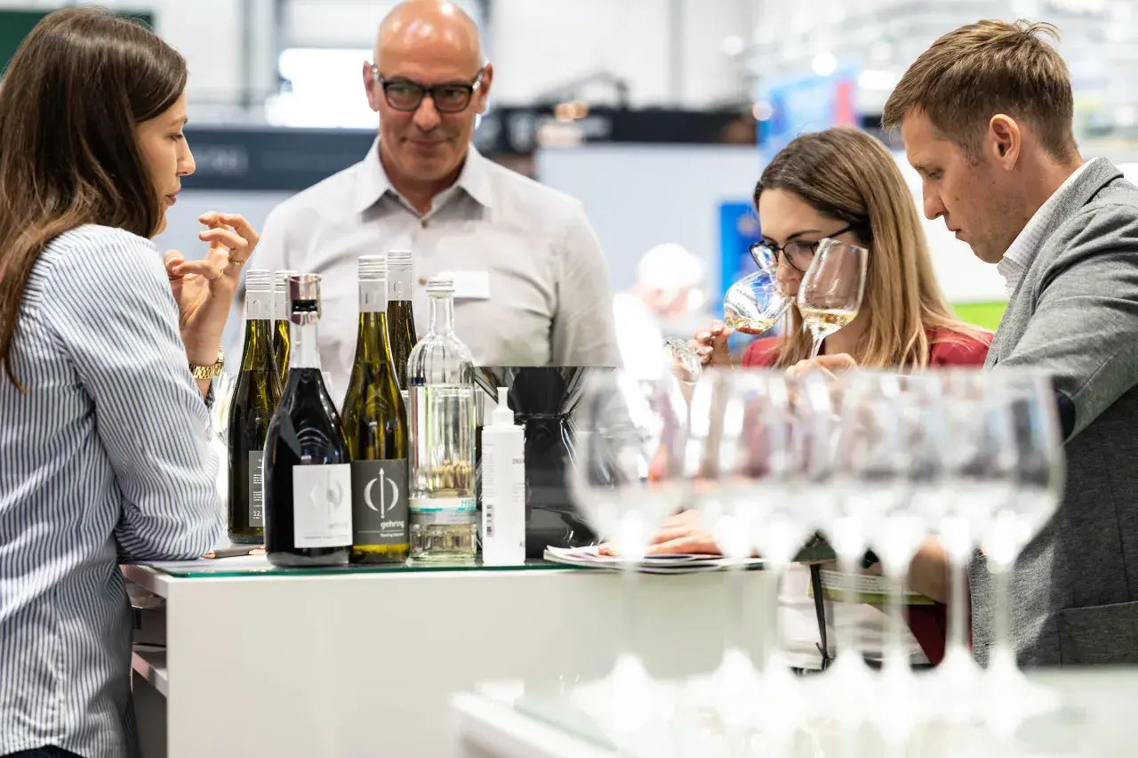 Rheinhessen Winzer präsentieren ihre Weine. Eine Frau verkostet ein Glas, während ein Mann und eine weitere Frau aufmerksam zusehen.
