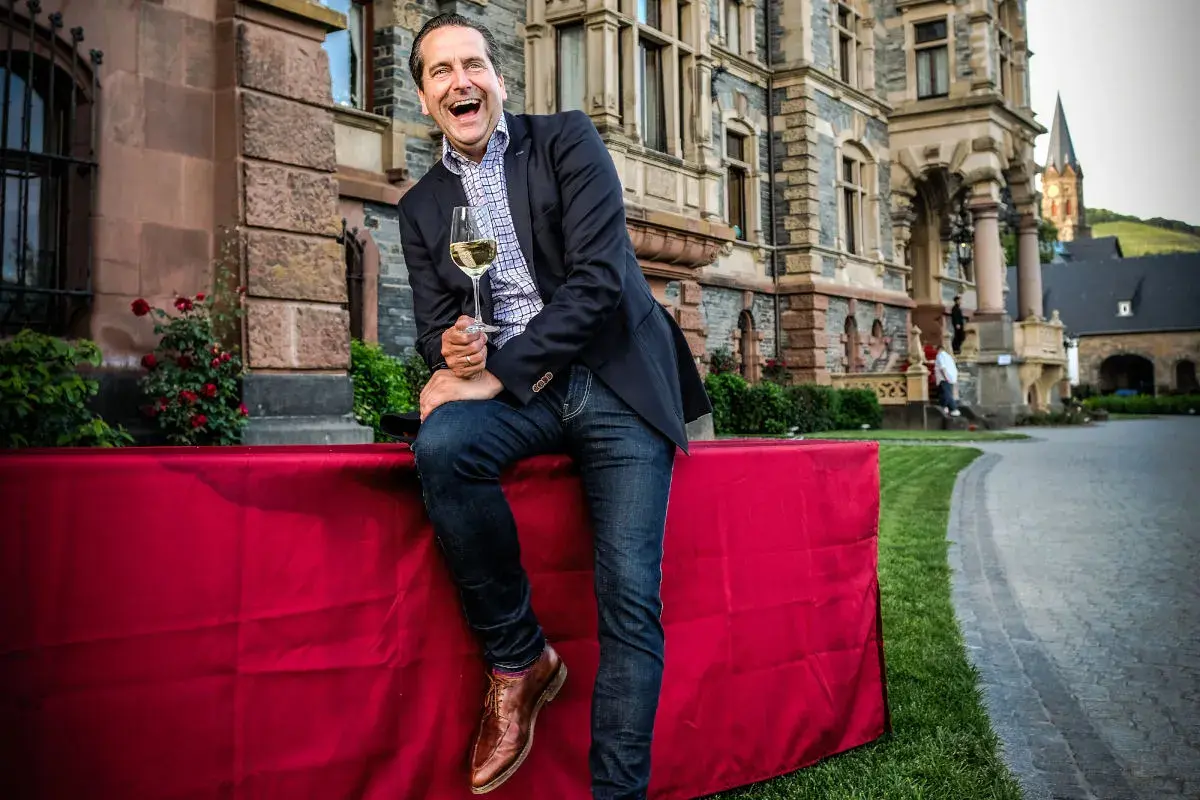 Gerhard Retter lacht ausgelassen mit einem Glas Wein in der Hand vor einem Schloss.