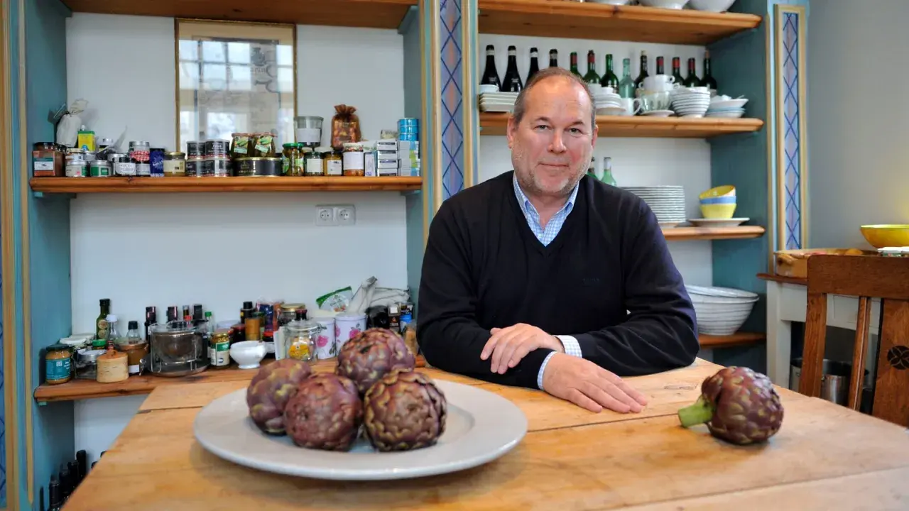 Otto Geisel sitzt an einem Holztisch mit Artischocken. Regale voller Gläser und Flaschen sind im Hintergrund zu sehen.