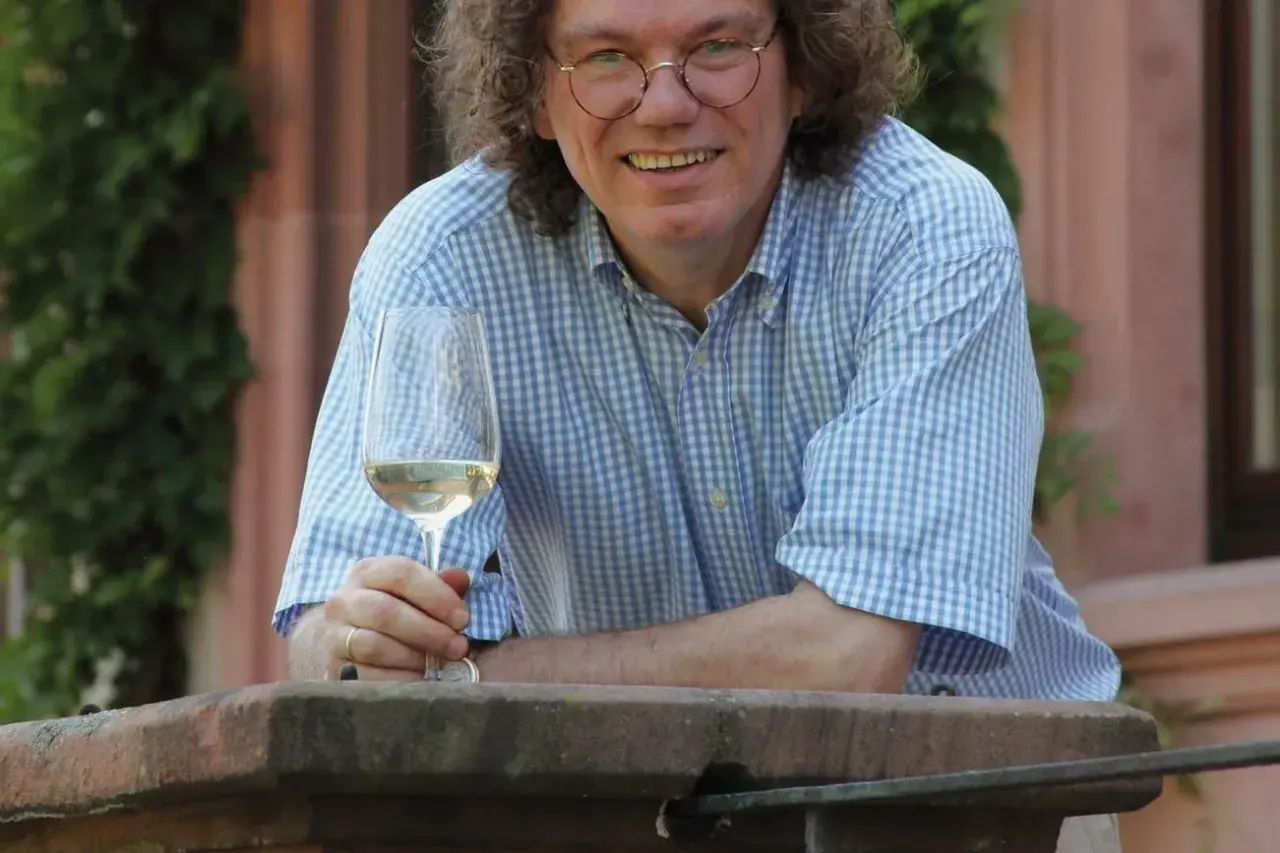 Ernst Loosen, mit lockigem Haar und Brille, hält ein Glas Weißwein. Er trägt ein blau-weiß kariertes Hemd und lächelt.