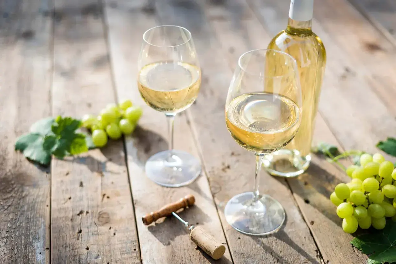 Zwei Gläser Weißwein, eine Flasche, Trauben und ein Korkenzieher auf Holztisch. Der Unterschied Riesling Chardonnay wird hier visuell angedeutet.