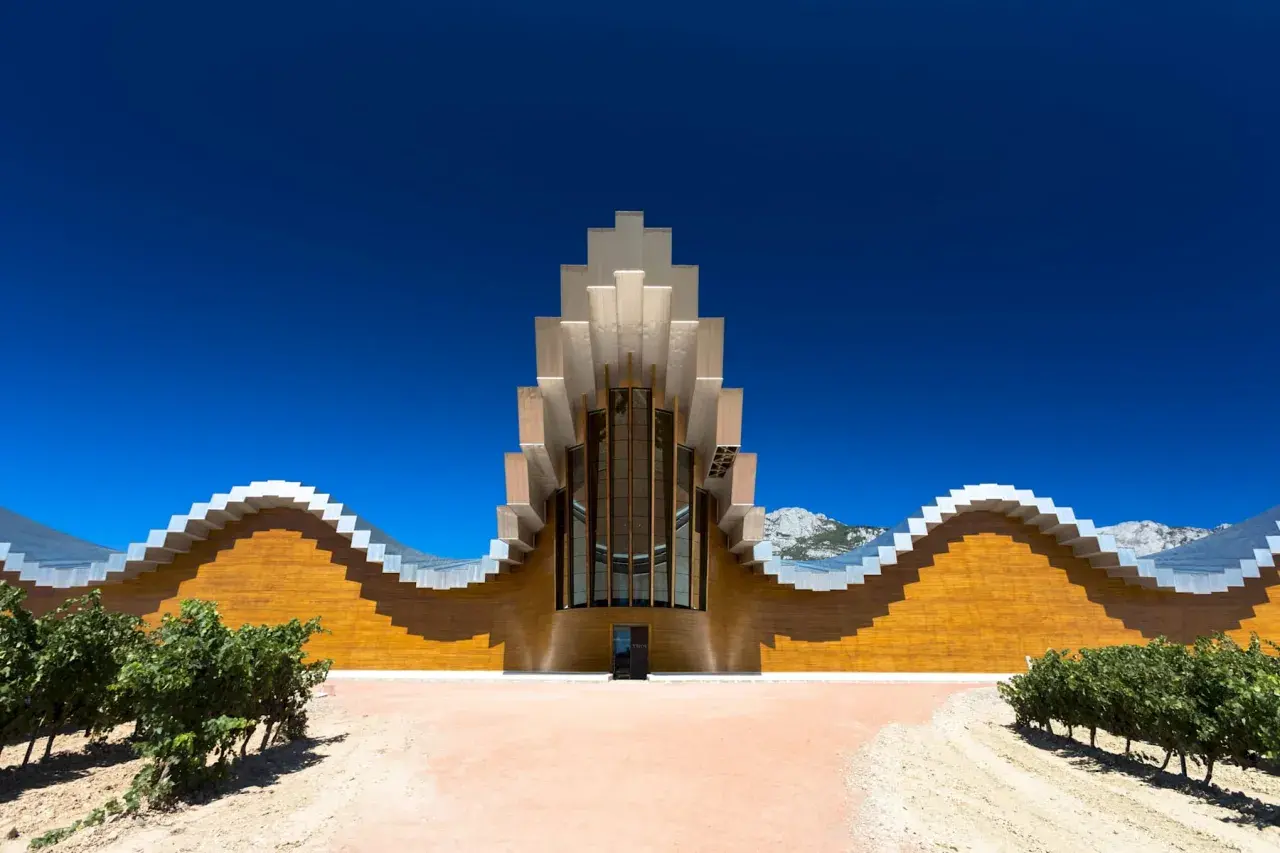 Moderne Weinbodega mit wellenförmiger Fassade und metallischem Dach in Rioja Alavesa, umgeben von Weinbergen und Bergen.