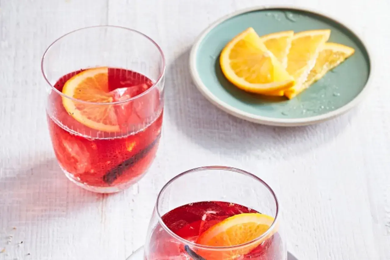 Zwei erfrischende Cocktails mit Portwein, garniert mit Orangenscheiben und Zimtstangen, stehen bereit. Daneben ein Teller mit frischen Orangenspalten.
