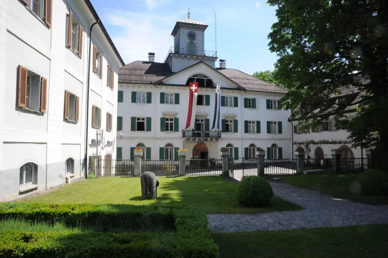 Das Schloss Reichenau mit Schweizer Fahnen und einem Uhrturm. Ein gepflegter Garten mit einem Stein und Büschen lädt zum Verweilen ein.