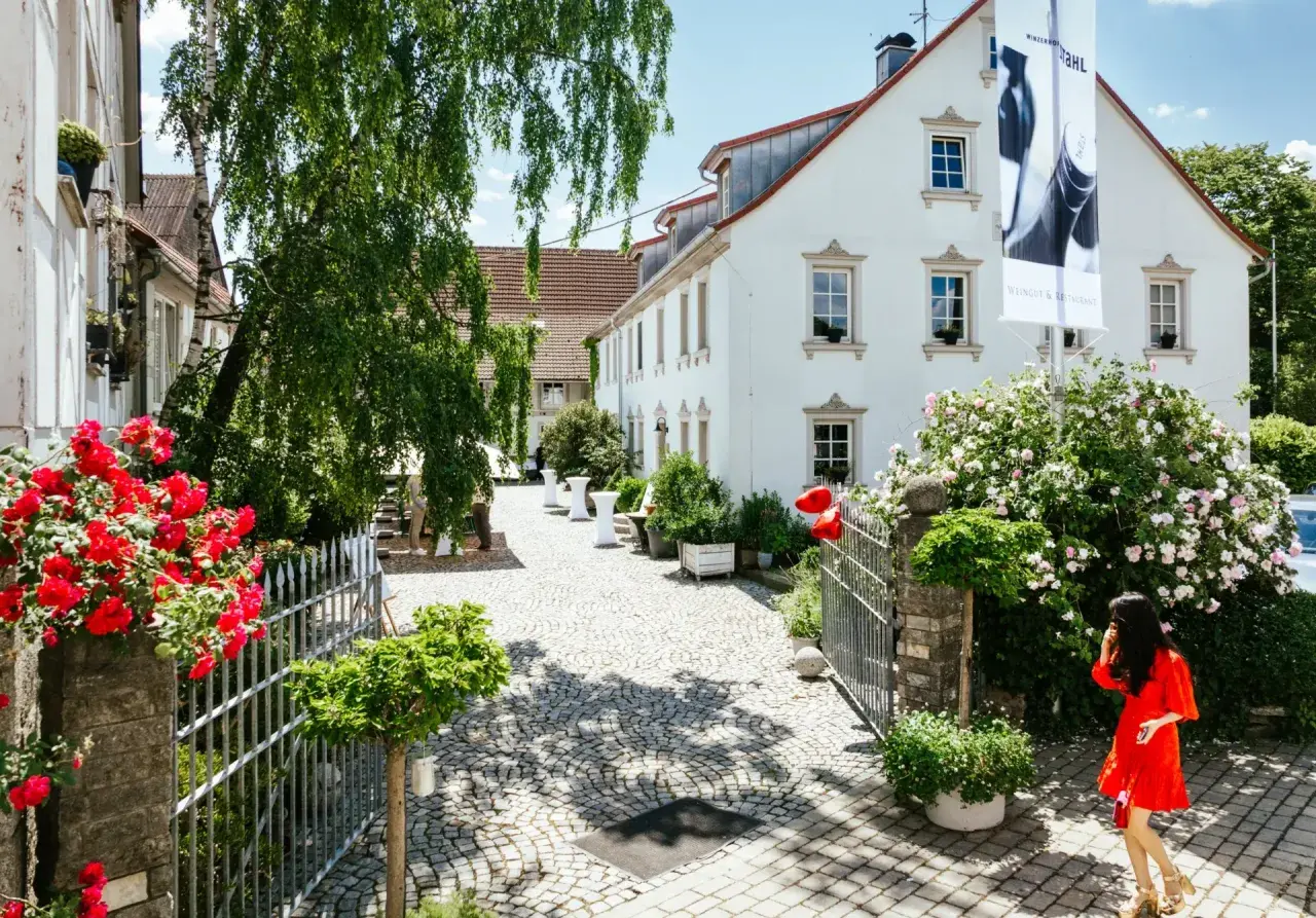Ein sonniger Tag am Weingut Christian Stahl. Rote Rosen blühen vor dem Tor, eine Frau im roten Kleid betritt das Anwesen.