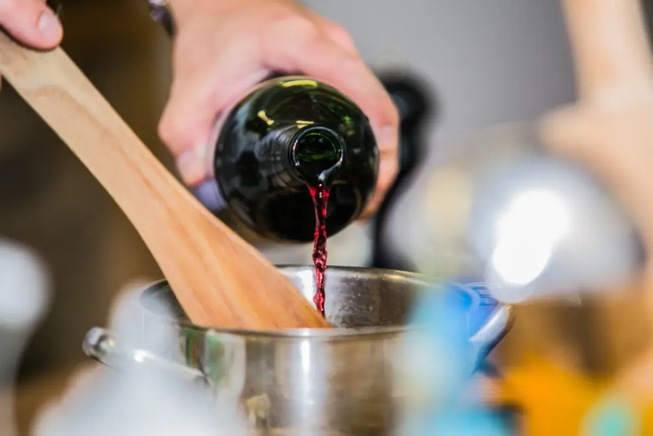 Rotwein wird in einen Topf gegossen, während ein Kochlöffel daneben liegt. Die Frage, wie lange braucht Rotwein zum Verkochen, ist hier relevant.