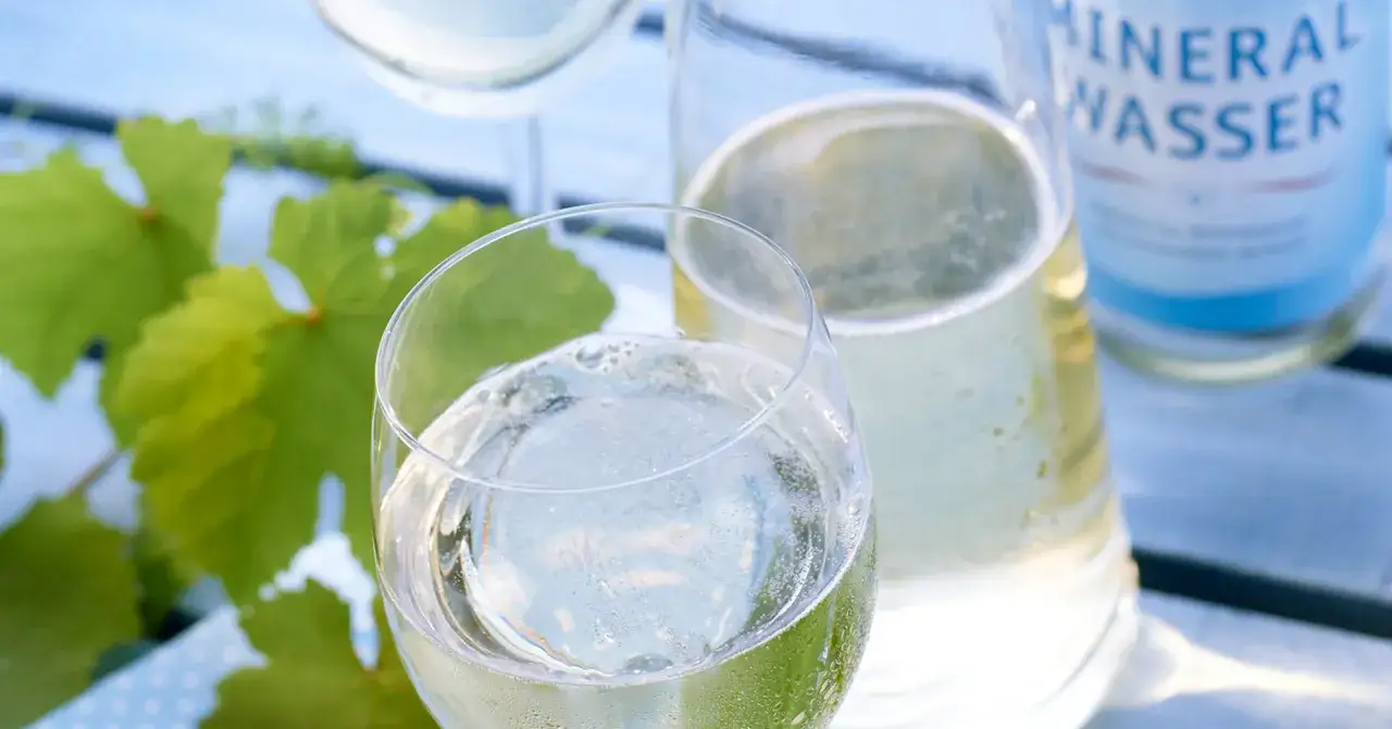 Erfrischende Weinschorle-Mischung im Glas, daneben eine Flasche Mineralwasser und gr&uuml;ne Weinbl&auml;tter.
