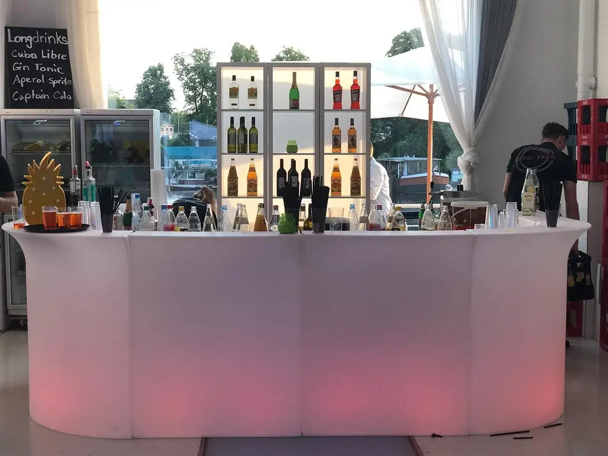 Leuchtender Tresen mit vielen Flaschen, bereit f&uuml;r Cocktails mit Gin. Ein Ananas-Dekor und Gl&auml;ser mit Getr&auml;nken stehen bereit.