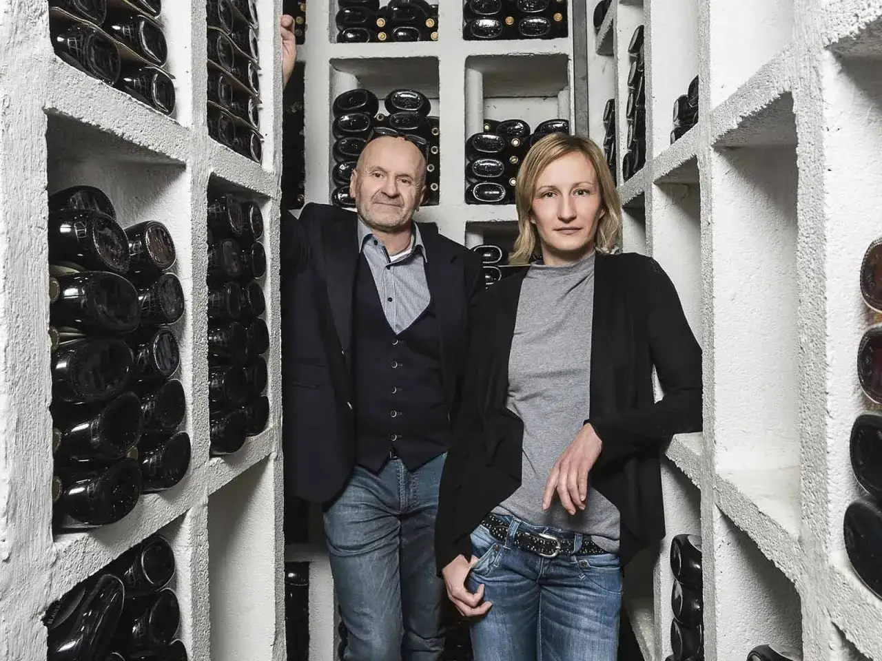Ein Mann und eine Frau stehen in einem Weinregal. Sie sind umgeben von vielen Flaschen Wein. Es ist wie es ist Wein.