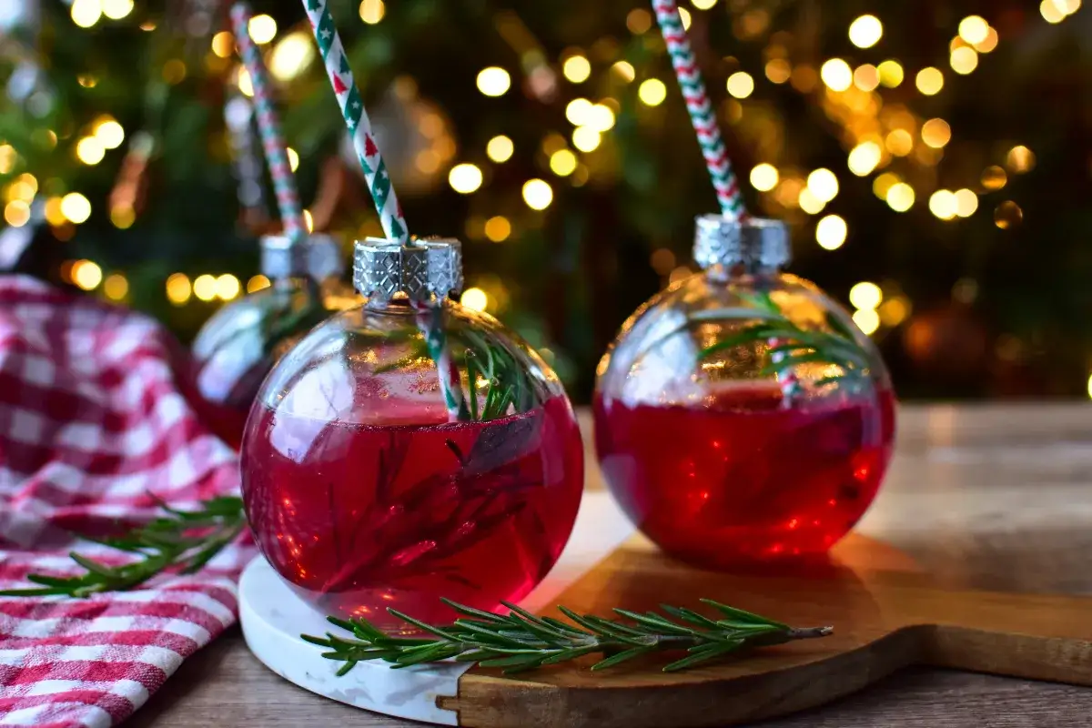 Zwei rote weihnachtscocktails mit Sekt in Glaskugeln, garniert mit Rosmarin, vor einem Weihnachtsbaum mit Lichtern.