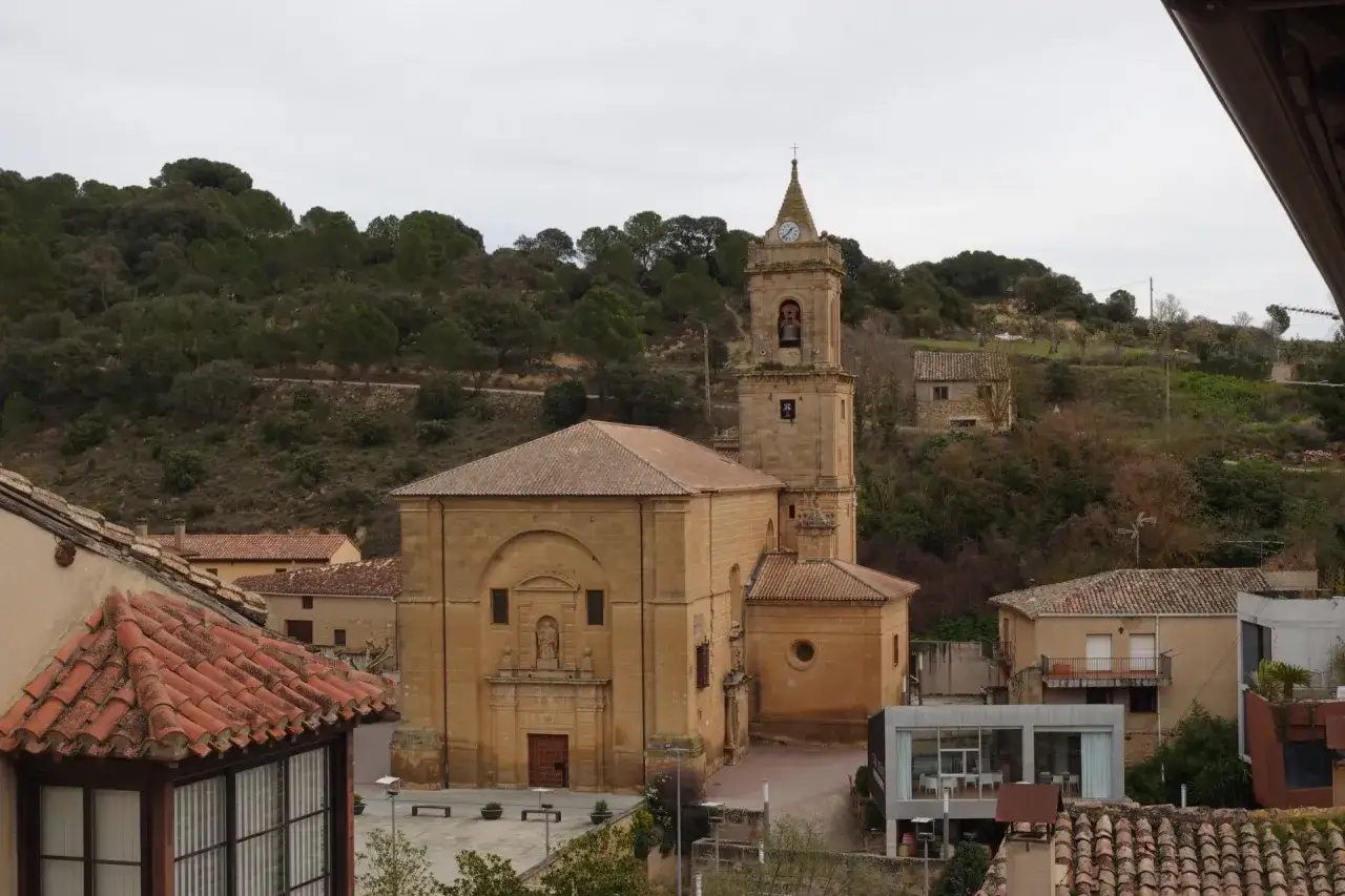 Kirche mit Glockenturm und Uhr in Rioja Alavesa, umgeben von H&auml;usern und bewaldeten H&uuml;geln.