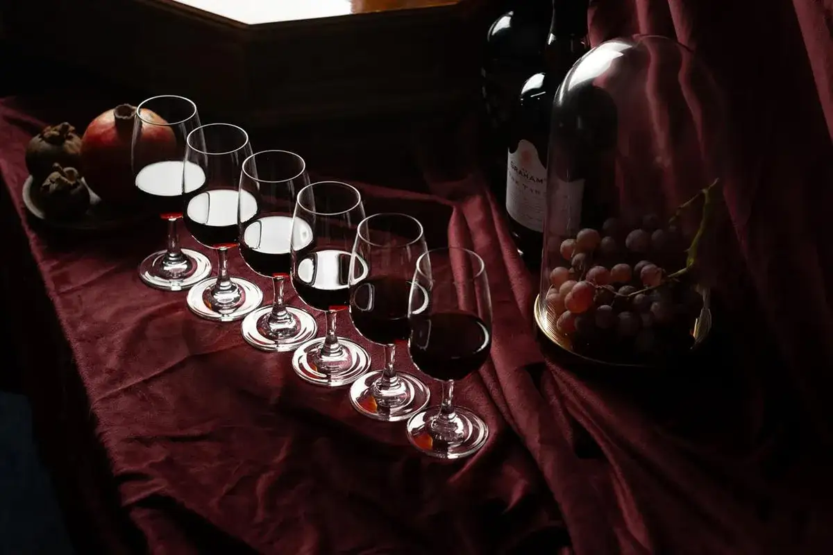 Sechs Gl&auml;ser mit dunklem Wein, bereit f&uuml;r Cocktails mit Portwein. Daneben Trauben unter einer Glasglocke und eine Flasche Portwein.