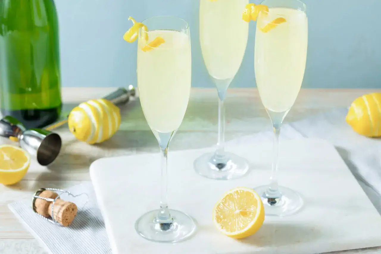 Drei elegante Gl&auml;ser mit einem erfrischenden Champagner Cocktail, garniert mit Zitronenzesten. Daneben liegen frische Zitronen und ein Korken.