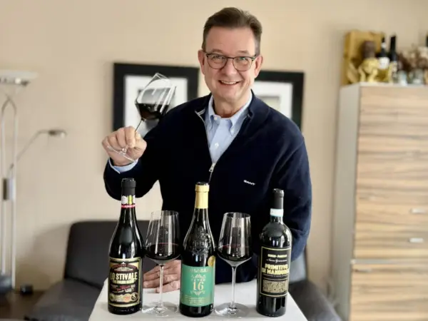 Luca Maroni Punkte: Mann mit Brille hält Weinglas, daneben drei Flaschen Rotwein und Gläser.